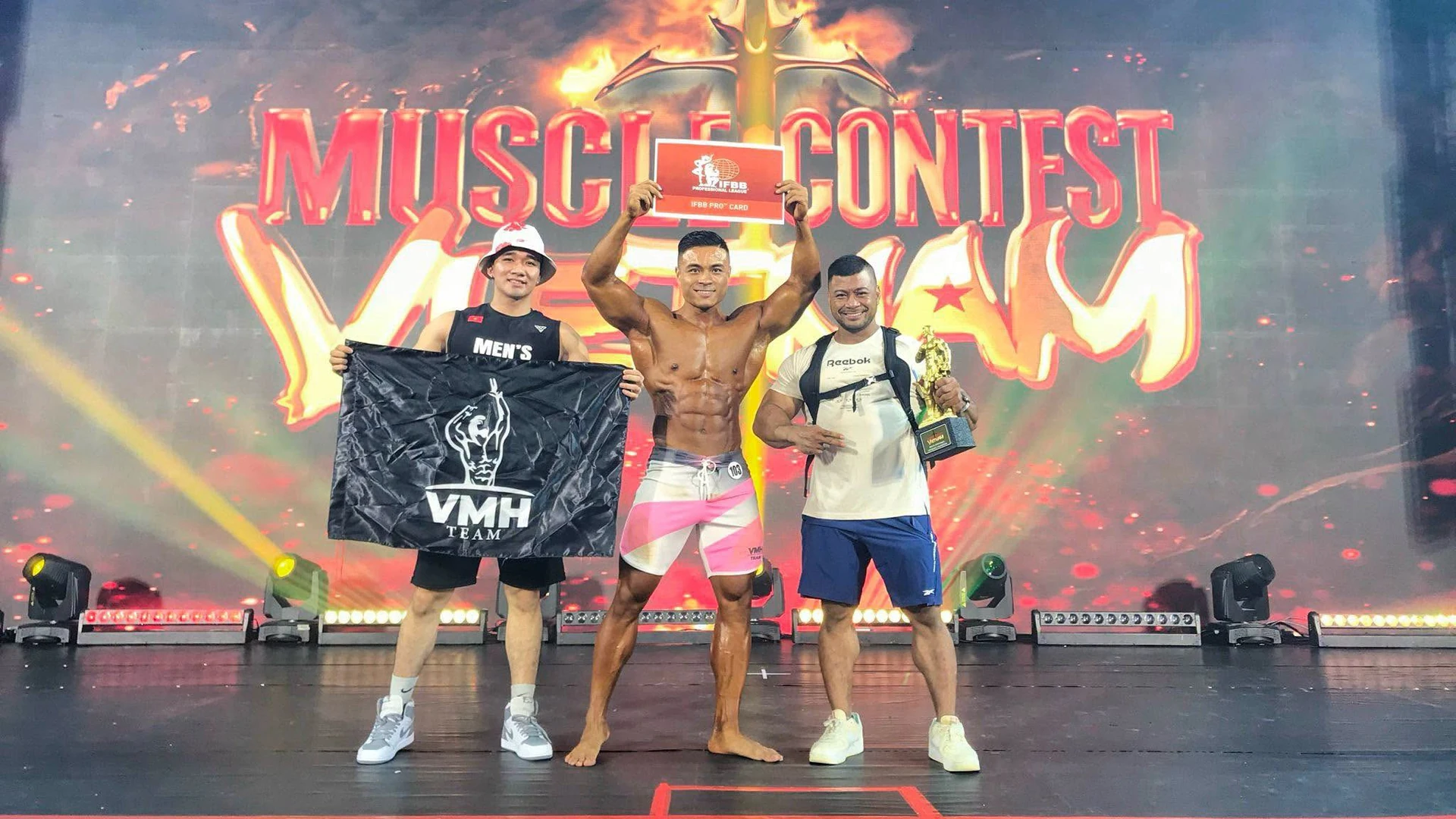 Vũ Minh Hiếu cùng hai người đồng hành, bảng IFBB Pro Card và lá cờ VMH Team trên sân khấu Musclecontest Vietnam